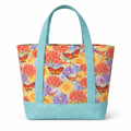 A structured cotton tote bag with a multicolor floral and butterfly print and aqua blue straps and base, featuring a front exterior pocket centered between vertical straps.