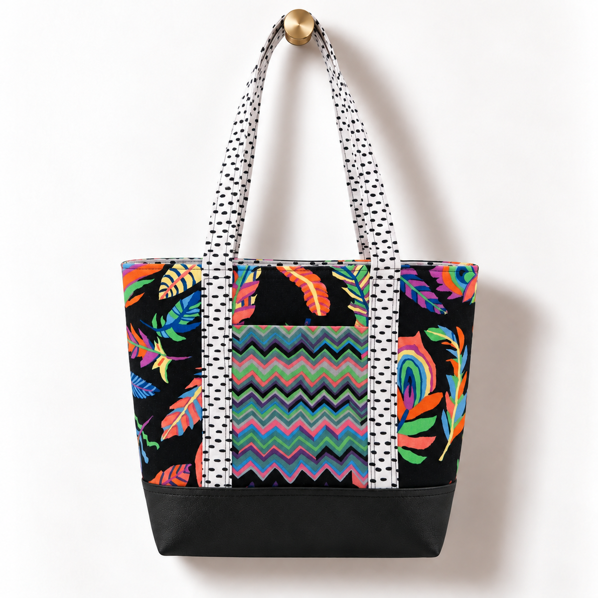Handmade cotton tote bag with colorful tropical feather pattern and white dotted straps hanging on a gold knob