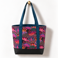 Handmade cotton tote bag with vibrant owl feather pattern in pink and teal tones, teal handles, and structured black base hanging on a gold knob