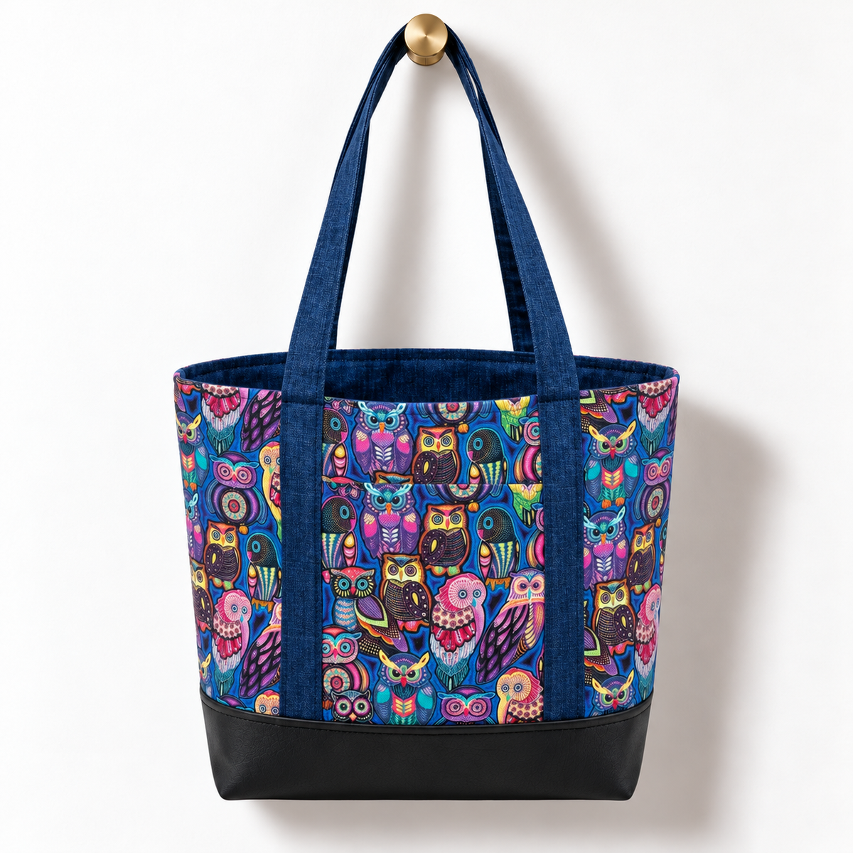 Handmade cotton tote bag with colorful owl pattern, navy handles, and structured black base hanging on a gold knob